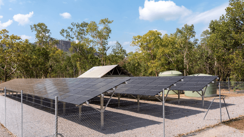 Solar panels in bushland in front of a metal roof building with a large plastic water tank behind to the right. .
