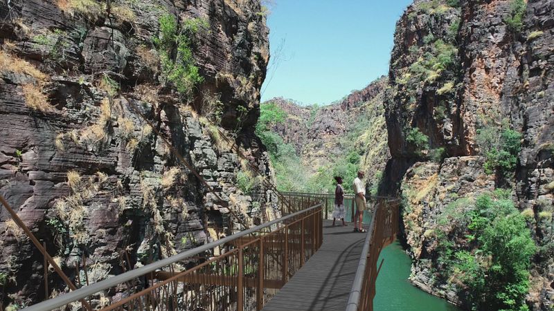 2 people walking along a metal walkway with a steel rail fence on both sides, which runs between 2 tall stone cliffs.