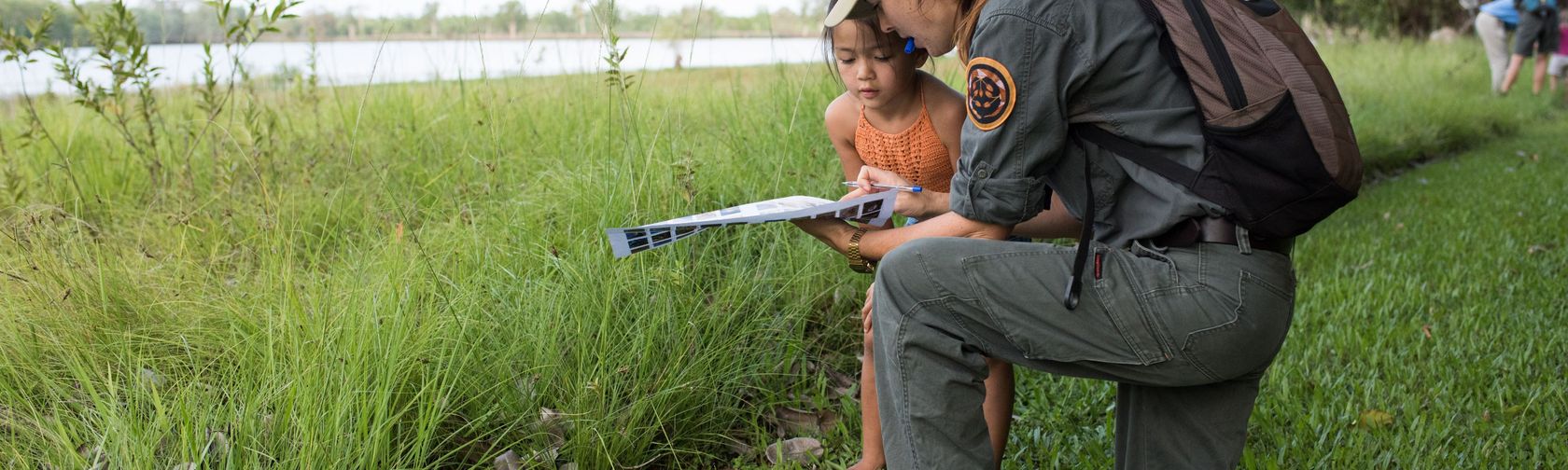 A female park ranger in a green shirt and long pants and wearing a backpack and cap, kneeling on a grassy river bank, showing a clipboard to a young girl in an orange dress.  
