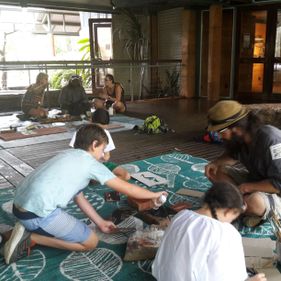Two children in white shirts and a man in a dark shirt and straw hat kneeling on the floor of Bowali Visitor Centre, painting on green paper with their hands and fingers. Behind them is 3 people sitting on the floor