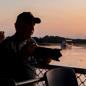 A man holding a camera and wearing a cap, standing on the deck of a boat looking out over the Yellow River sunset